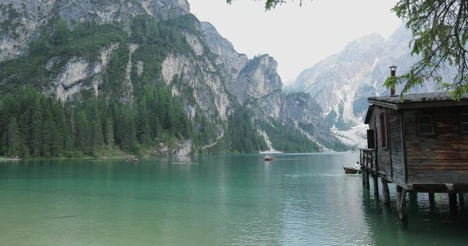 smooth ride from a steadicam because of the house and tree branches to Lake Braies against the backdrop of rocky mountains and floating kayak boats on the water. overall plan