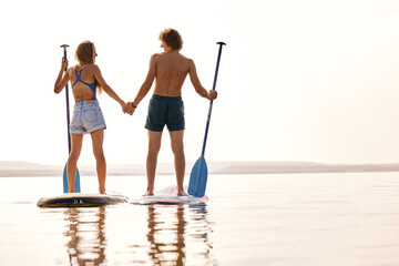 View from back on fit couple standing on surfboard, holding hands together, man and woman in love....
