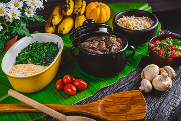 FEIJOADA VEGETARIANA DE COGUMELOS NA CUMBUCA PEQUENA COM ACOMPANHAMENTOS ARROZ, COUVE, FAROFA, BANANA DECORADA COM FOLHA DE BANANEIRA E FLORES