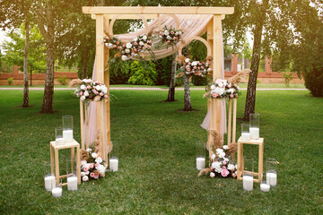 A beautiful wooden wedding arch decorated with flowers and candles stands on the green grass in the...