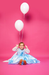 Obraz na płótnie Canvas A girl in a blue dress sits on a pink background decorated with white balloons. Cute baby smiles and grimaces. Vertical photo of a happy child on his birthday. Children's holiday concept