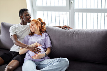 diverse young man and woman relax on comfortable couch sofa, waiting baby. pregnant lady with husband dream of bright happy future together, hug cuddle on sofa in living room enjoy weekend