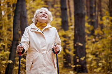 European aged lady with gray short hair standing in forest with Nordic walking sticks, going home...