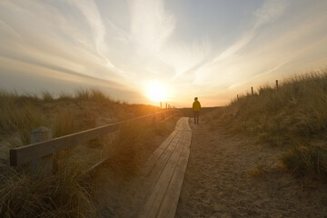 Eine Person geht auf einem Holzbohlenweg in einer Dünenlandschaft am Meer, und die Sonne ist...
