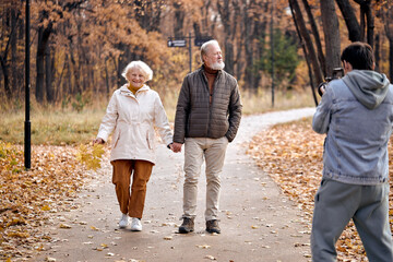 Fototapeta premium Aged active caucasian couple holding hands looks happy in park in autumn season,happy anniversary concept. gray haired mature man and woman in coats walking together, in love. backstage of photoshoot