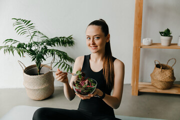 Attractive woman eating bowl of healthy salad after working out at home, sitting on a yoga mat. Healthy life concept