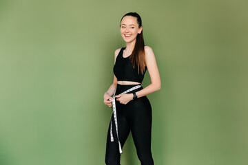 Laughing young brunette woman measuring her thin waist with a tape measure, on green background. Weight loss diet results concept