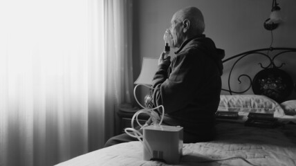 Black and white back view angle of an old man with asthma using a nebulizer in his room sitting on a bed - obrazy, fototapety, plakaty