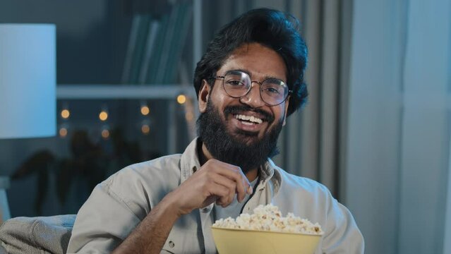 Portrait arabic hispanic latino indian muslim bearded man happy relaxed guy in glasses watching movie program funny series tv online eating popcorn laughing having fun at home in evening night dark