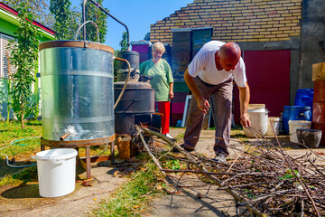 Woman is manually mixing fruit marc in distillation apparatus, man is throwing firewood for...
