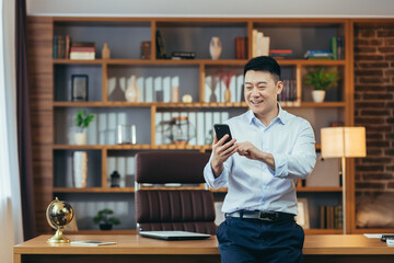 Successful Asian businessman in modern home office, looks at phone screen, smiles and rejoices,...