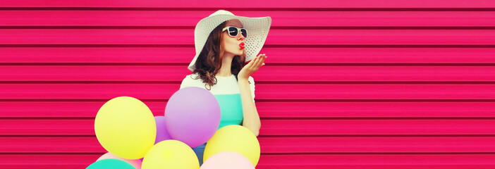 Portrait of beautiful young woman with bunch of balloons blowing her lips sends air kiss wearing a colorful dress on pink background