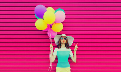 Portrait of beautiful young woman with bunch of balloons blowing her lips sends air kiss wearing a colorful dress on pink background
