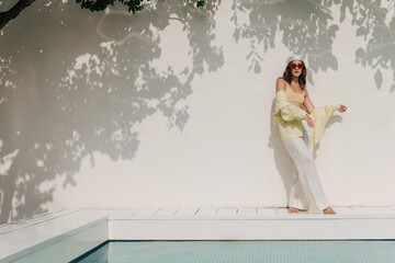 Full-length young brunette caucasian girl in sunglasses stands against white wall on warm day....