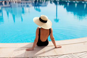 a girl in a swimsuit and a large hat view from the back. A woman in a black one-piece swimsuit sits on the edge of the pool with her back to the camera.