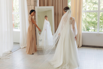 Side view of two happy smiling girls preparing for wedding in the morning. The young bride stands...