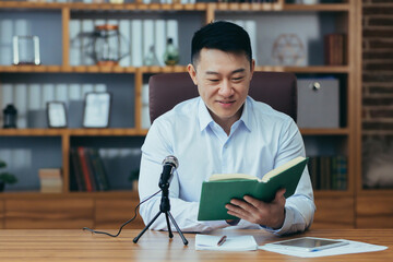 Asian teacher records online business course, uses microphone, records audio podcast, and reads...