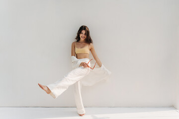 Full length funny young caucasian woman raising leg in front of her while standing against white background. Brunette with wavy hair smiles and wears top, pants. Waiting, loneliness and beauty concept