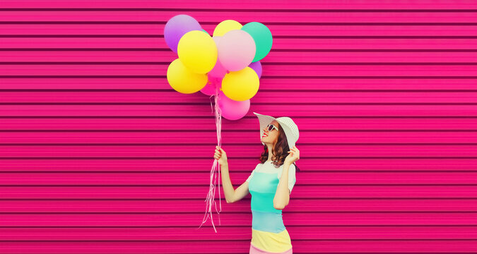 Portrait of beautiful happy smiling young woman with bunch of balloons wearing a colorful dress on pink background