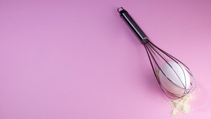 Whisk with an egg inside, flour lies next to it. Pink background with space for text. Picture for...