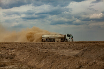 Tank truck and yellow dust cloud on sand road against grey cloudy sky. Road construction site.