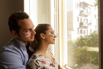 coppia di giovani che con i colori de dell'Ucraina disegnati sul viso, guardano fuori dalla finestra di casa loro - obrazy, fototapety, plakaty