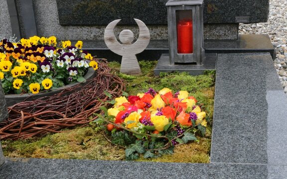 Mit Blumen geschmückte Gräber an einem katholischen Friedhof in Österreich, ein Blumengebinde mit Rosen, daneben eine Blumenschale mit Stiefmütterchen und ein Engel und eine Friedhofslaterne