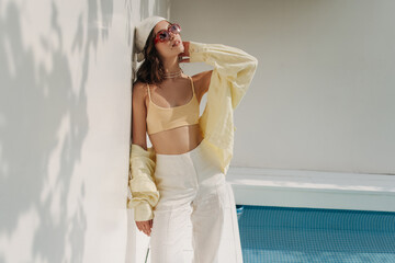 Relaxed young caucasian woman basking in sun while standing against white wall outdoors. Pretty...