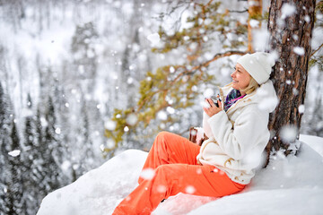 excited blonde caucasian female sit on top of mountains drinking hot tea from thermos, enjoying the landscape, looking at side in contemplation, at frozen snowe winter day, in nature