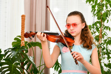 A girl in sunglasses plays the violin at home among green plants.