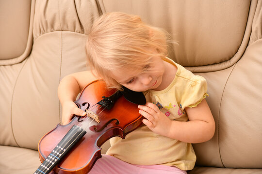 A little girl sits on a sofa with a violin and does not know what to do with it.