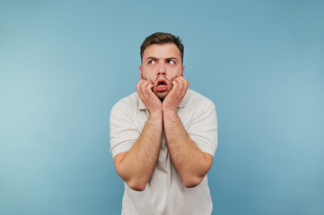 Shocked young man with bristles clings to his head and looks away with a surprised face on a blue background.