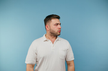 An adult man with bristles in a white T-shirt stands on a blue background and looks away with a serious face.