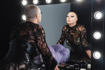 man in bright makeup and black lace clothing looking in mirror in dressing room.