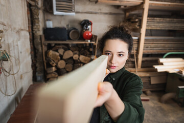 Brunette carpenter checking wooden plank in blurred workshop.