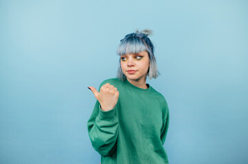 Beautiful girl with blue hair and a serious face stands on a blue background and looks and points to the side on copy space - obrazy, fototapety, plakaty