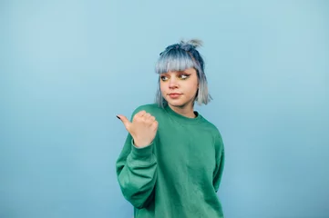 Fotobehang Beautiful girl with blue hair and a serious face stands on a blue background and looks and points to the side on copy space © bodnarphoto