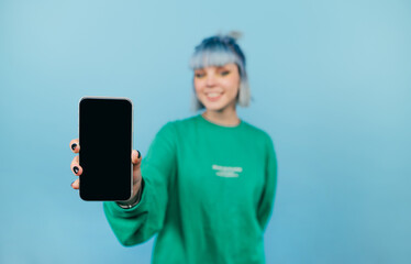 Positive hipster girl with blue hair and with a smile on her face shows to the camera a smartphone...