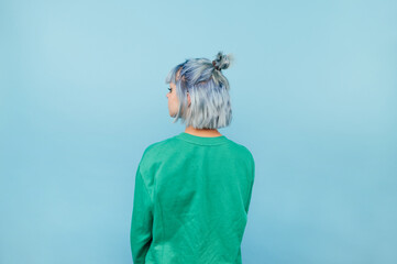 Hipster girl with blue hair stands with her back to the camera on a blue background and looks away,...