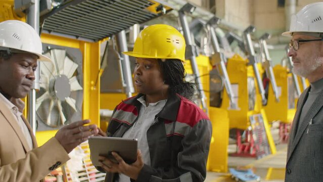 African American female engineer in hardhat using digital tablet and speaking with multiethnic managers while working in machine factory