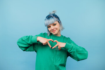 Positive hipster girl with colored hair in a green sweatshirt stands on a blue background and shows a gesture of love to the camera