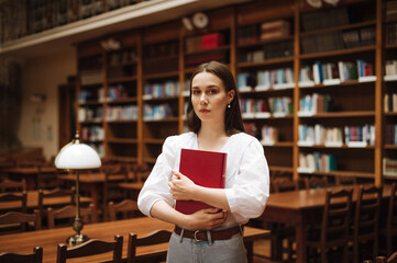 Beautiful woman in a white blouse stands with an atmospheric cozy library with a book in her hands and poses for the camera with a serious face.