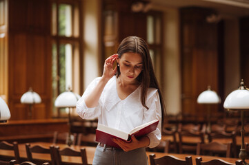 Attractive woman in blouse reads a book standing in the library with a serious face.