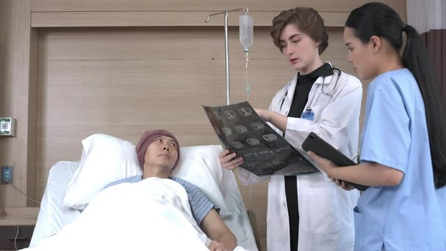 A young short-haired Caucasian female doctor in a white gown diagnosing patient's brain MRI scan film to Asian female therapist holding a tablet, with Asian male cancer patient lying listening on bed.