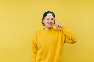 Positive young man in a cap stands on a yellow background, shows a finger on his temple showing a gesture of 
