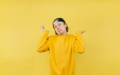 Funny scared guy in a cap on a yellow background looks puzzled at the camera and spreads his arms.