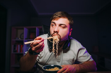 Funny man eating vermicelli with hungry face for dinner while working on the computer in a dark room at home.