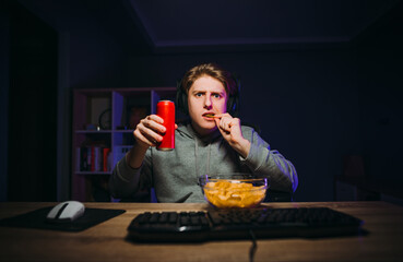 A hungry gamer in a headset eats chips and drinks sweet water with a serious face looking at a computer screen during a night of play.