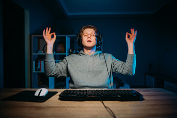 The guy gamer meditates sitting at night at the computer in a headset, closed his eyes and with his hands raised soothes himself with yoga. © bodnarphoto