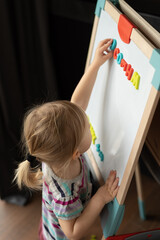 a little fair-haired girl stands near a magnetic board on a children's easel. 